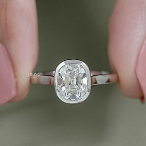 May include: A silver ring with a large, cushion-cut diamond. The diamond is set in a bezel setting, and the band is thin and polished. The ring is held between two fingers with pink nail polish.