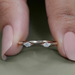 Puede incluir: Un anillo de oro rosa con dos diamantes talla marquesa a cada lado de un diseño abierto. El anillo se sostiene entre dos dedos, resaltando su estilo delicado y elegante. Los diamantes brillan sobre el metal cálido.