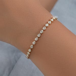 May include: A gold bracelet adorned with a sequence of pear-shaped, clear gemstones. The bracelet is displayed on a wrist, highlighting the elegant design and sparkle of the gemstones. The bracelet is a piece of jewellery.