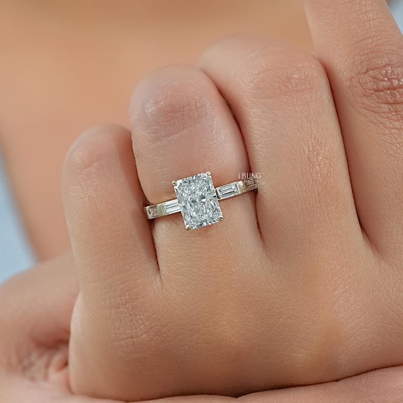May include: A radiant-cut diamond engagement ring with a gold band. The ring features a large rectangular center stone and smaller baguette diamonds on the sides. The ring is on a person's finger.
