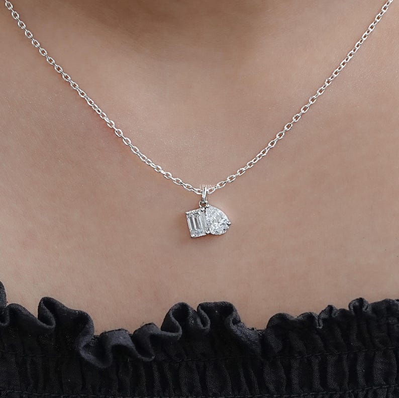 May include: A delicate silver necklace featuring a pendant with two diamond-cut gemstones. One is an emerald cut, and the other is a pear shape. The necklace is displayed against a neutral background.