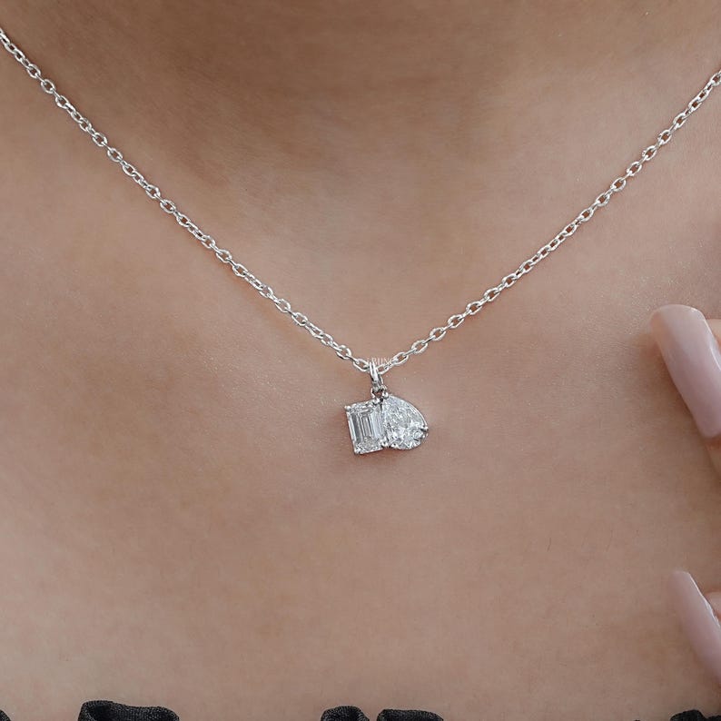 May include: A delicate silver necklace featuring a pendant with two gemstones: an emerald-cut and a pear-shaped diamond. The necklace is worn around the neck, showcasing the sparkling pendant.