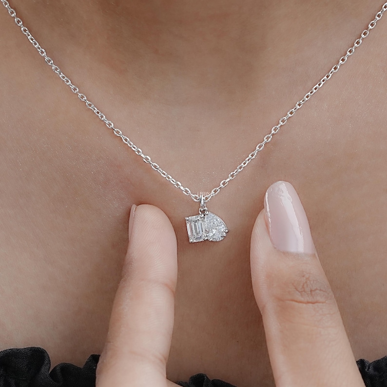 May include: A delicate silver necklace featuring a pendant with two sparkling gemstones: a rectangular emerald cut and a pear-shaped diamond. The necklace is displayed against a neutral background.