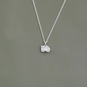 May include: A delicate silver necklace featuring two clear, sparkling gemstone pendants: an emerald cut and a pear shape. The necklace hangs against a neutral gray background, highlighting the pendant's brilliance. A simple and elegant piece of jewelry.