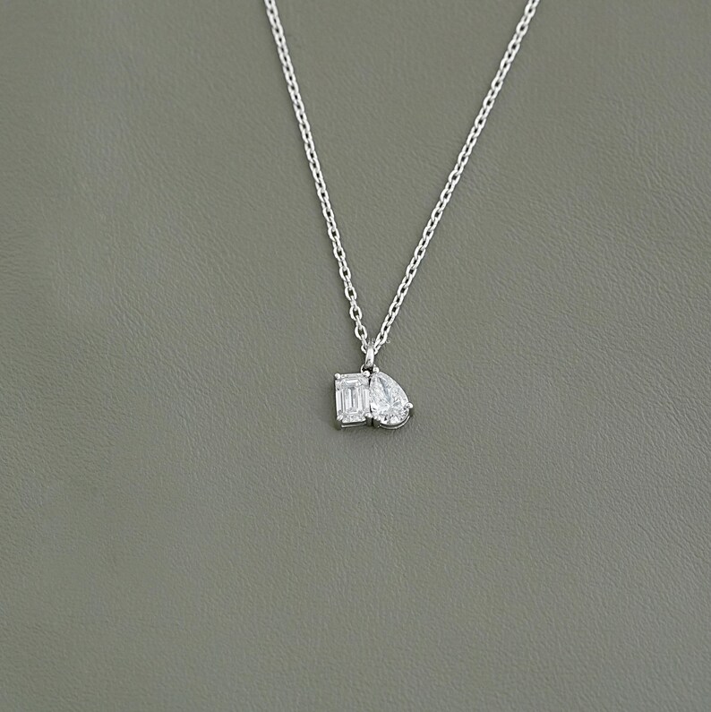 May include: A delicate silver necklace featuring two clear, sparkling gemstone pendants: an emerald cut and a pear shape. The necklace hangs against a neutral gray background, highlighting the pendant's brilliance. A simple and elegant piece of jewelry.