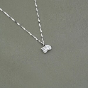May include: A delicate silver necklace featuring a pendant with two white gemstones: an emerald-cut and a pear-shaped stone. The necklace has a fine chain and is set against a gray background.