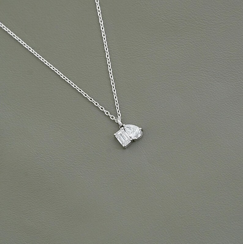 May include: A delicate silver necklace featuring a pendant with two white gemstones: an emerald-cut and a pear-shaped stone. The necklace has a fine chain and is set against a gray background.