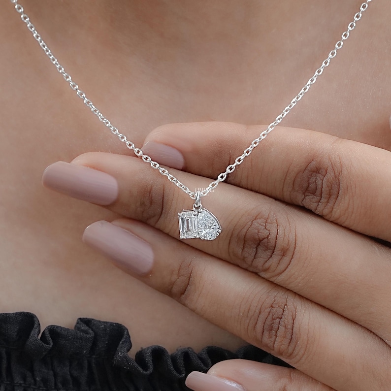 May include: A delicate silver necklace featuring a pendant with two white gemstones. One gemstone is an emerald cut, and the other is a pear shape. The necklace is displayed on a person's hand.