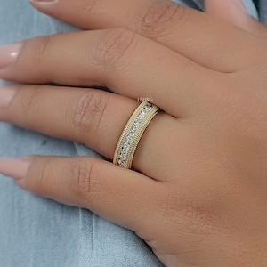May include: A gold band ring with a textured outer edge and a row of small, clear stones set in the center. The ring is on a person's finger, with a light blue background.