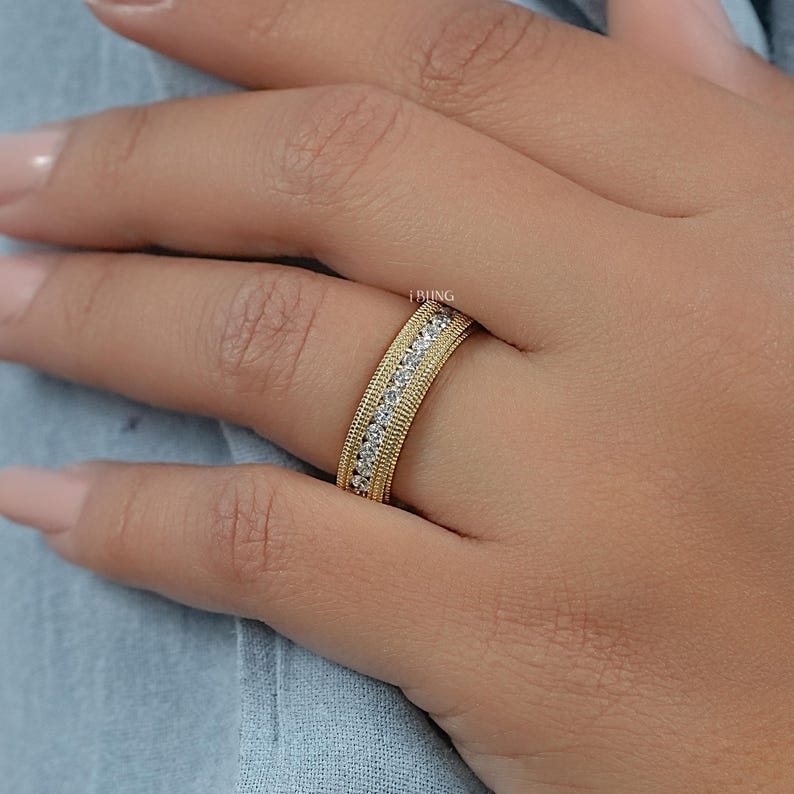 May include: A gold band ring with a textured outer edge and a row of small, clear stones set in the center. The ring is on a person's finger, with a light blue background.