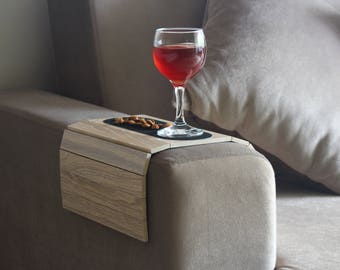 May include: A wooden armrest tray with a black surface for a glass of red wine and a small bowl of almonds. The tray is attached to a brown couch.