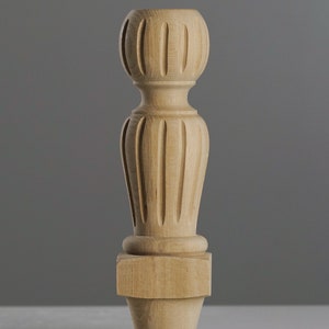 May include: A light-coloured, unfinished wooden furniture leg. The leg features a decorative, carved design with a rounded top and a square base. It measures 25 cm (9.85 inches) in height and 6.5 cm (2.56 inches) in diameter.