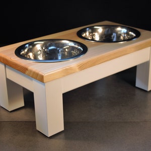 May include: A raised dog feeder with two stainless steel bowls set into a light-coloured wooden top. The top is supported by a white, rectangular base. The feeder is designed to elevate the bowls for easier access.