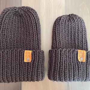 May include: Two dark brown knit beanies with a ribbed texture and a folded cuff. Each beanie features a small, rectangular tan leather tag attached with a silver metal rivet. The left tag has the text "da da" and the right tag has "mi ni".