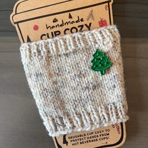May include: A cream-colored, speckled knit cup cozy featuring a green Christmas tree-shaped button. The cozy is displayed on a cardboard sleeve labeled "handmade A CUP COZY" and "REUSABLE CUP COZY TO PROTECT HANDS FROM HOT BEVERAGE CUPS!"