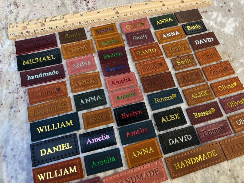 May include: A collection of leather labels with various names and the words "handmade" and "Jones" embroidered in gold thread. The labels are arranged in rows on a light brown surface.