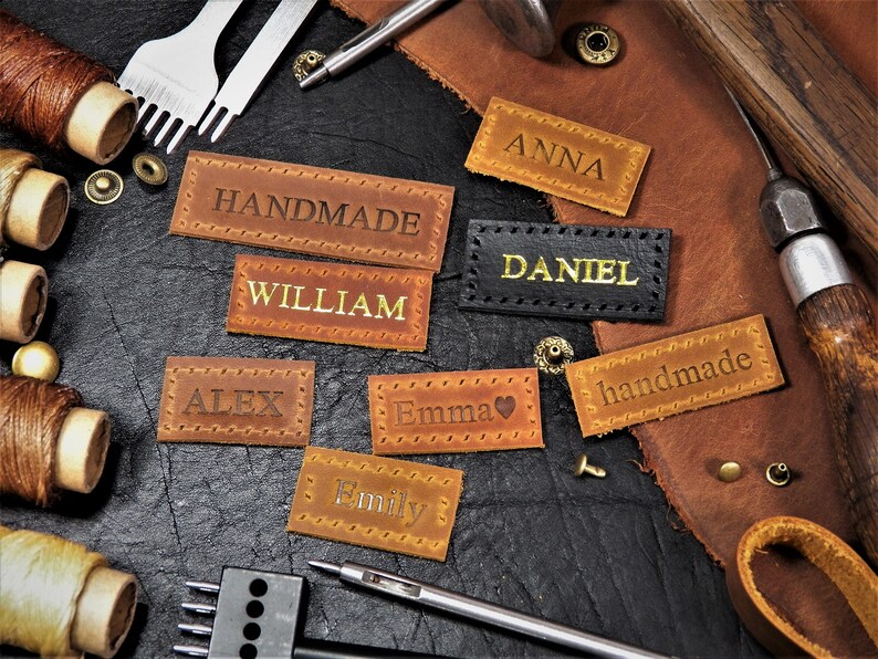 May include: Assortment of brown leather labels with various names and the words "handmade" and "William" in gold lettering. The labels are on a dark brown surface with leather working tools.
