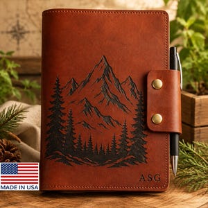 May include: A brown leather journal cover featuring an engraved mountain and pine tree landscape. The cover includes a side strap with two brass-colored metal snaps, a black pen inserted into the loop, and the initials ASG embossed at the bottom.
