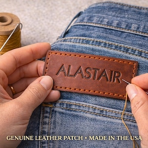 Op de afbeelding: Een bruin leren patch met de naam "ALASTAIR" in reliëf wordt op een blauwe spijkerbroek genaaid. Een spoel met gele draad en een naald zijn ook zichtbaar. De patch heeft een gestikte rand.