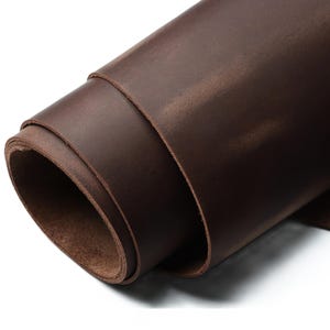 Pode incluir: Folha enrolada de couro castanho escuro. O couro tem uma superfície lisa, ligeiramente texturizada, com variações de cor, criando uma aparência natural e rústica. As bordas do rolo revelam a espessura e a textura do material.