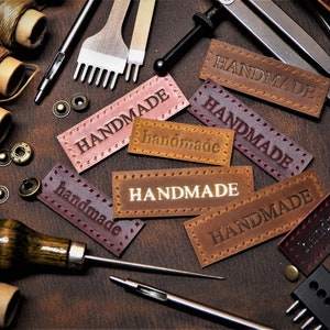 May include: A close-up of leather craft supplies, including spools of thread, leather punches, and leather labels that say "HANDMADE".