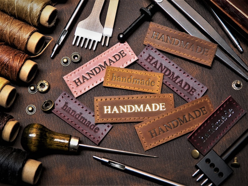 May include: A close-up of leather craft supplies, including spools of thread, leather punches, and leather labels that say "HANDMADE".