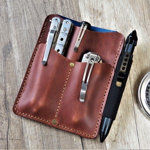 May include: A brown leather multi-tool organizer with multiple pockets, holding a multi-tool, pens, and a knife. A black pen and a compass are next to the organizer. The organizer is on a wooden surface.
