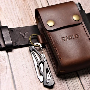 以下が含まれることがあります： 銀色のバックルが付いた茶色の革製のベルトと、茶色の革製のポーチが付いています。ポーチには「PAOLO」という名前が刻印されています。ポーチは金属製のクリップでベルトに取り付けられており、前面に小さなポケットがあります。黒いマルチツールはキーリングでベルトに取り付けられています。