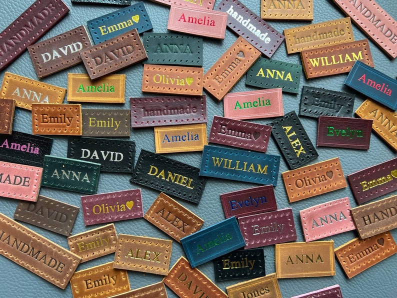 May include: A collection of brown leather labels with various names printed in gold, white, and black lettering. The labels have a stitched border and some have a small heart symbol. The names include Amelia, David, Emma, William, Olivia, Alex, Daniel, Emily, and Anna.