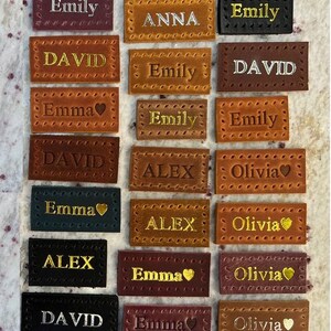 May include: A collection of 18 leather labels with various names, including Olivia, Anna, David, Emily, Emma, and Alex. The labels are brown, black, red, and green with gold lettering. Some labels have a small heart symbol next to the name. The labels are handmade and have a decorative stitched border.