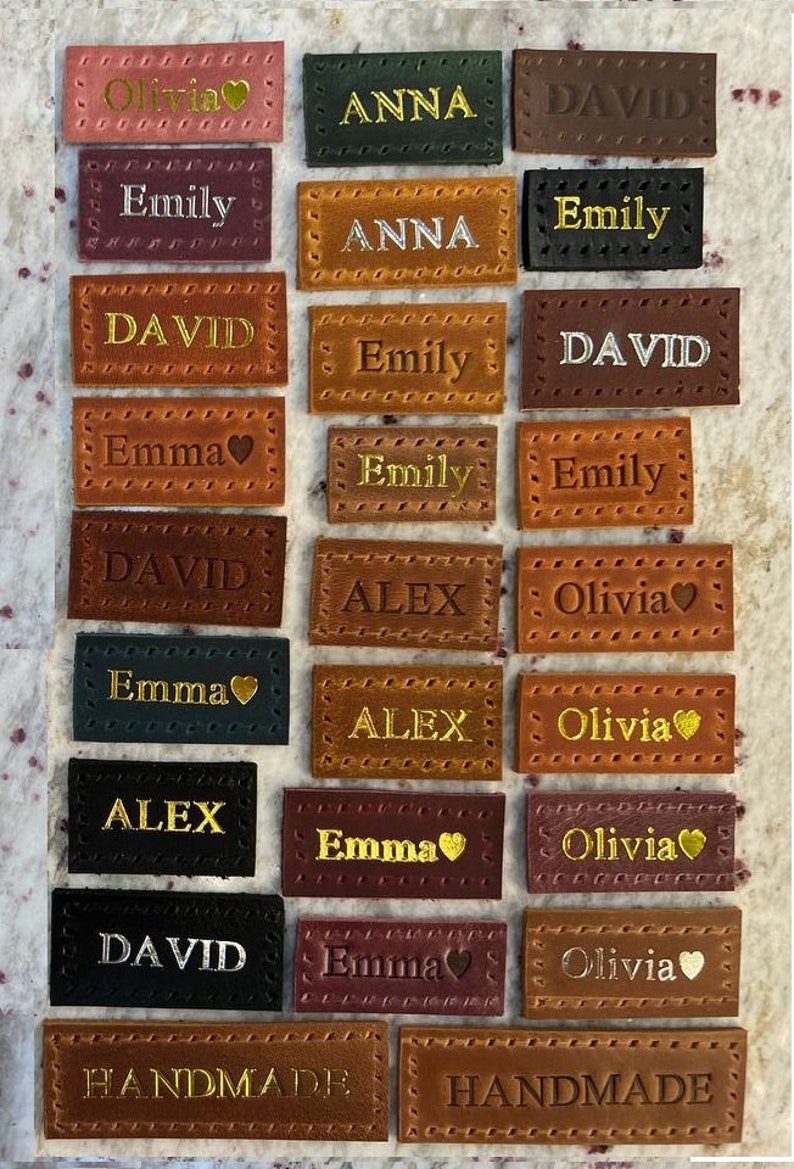May include: A collection of 18 leather labels with various names, including Olivia, Anna, David, Emily, Emma, and Alex. The labels are brown, black, red, and green with gold lettering. Some labels have a small heart symbol next to the name. The labels are handmade and have a decorative stitched border.
