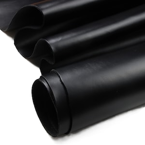 May include: Close-up of a stack of black leather sheets. The leather has a smooth, shiny surface and is folded and rolled, revealing the texture and depth of the material. The image highlights the rich, dark color and the supple nature of the leather.