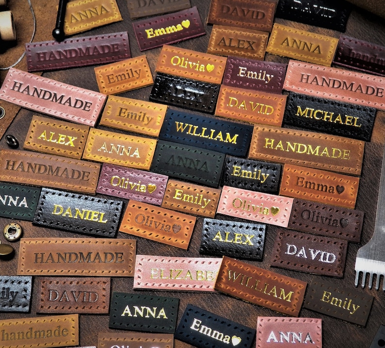 May include: A collection of brown leather labels with various names and the word "Handmade" printed on them. Some labels have a heart symbol next to the name.