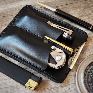 May include: Black leather pen and knife holder with a pen, knife, and lighter. The holder is on a wooden surface with a compass.