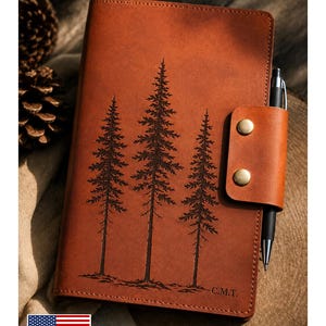 May include: A tan leather journal cover featuring a laser-engraved design of three pine trees and the initials C.M.T. in the bottom right corner. The cover includes a side strap with two brass-colored metal snaps and a black pen inserted.