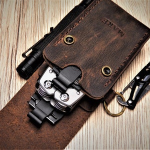 Może przedstawiać: Brązowy skórzany portfel z wielofunkcyjnym narzędziem i brelokiem. Portfel ma wygrawerowane słowo "MARE" na przodzie.