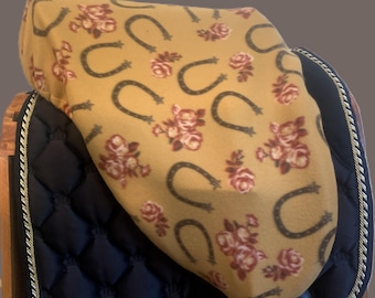 May include: A yellow fleece saddle pad with a pattern of brown horseshoes and pink roses. The saddle pad is folded over a black saddle pad with a gold braid trim.