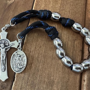 Op de afbeelding: Een zilveren rozenkrans met een kruisbeeld en een St. Michael-medaille. De rozenkrans kralen zijn zilver, en het koord is zwart met blauwe accenten. De achtergrond is een houten oppervlak.