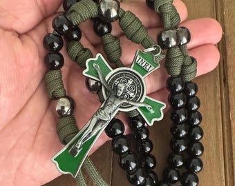 Puede incluir: Un rosario negro y verde con una medalla de San Benito y un crucifijo. El rosario está hecho de cuentas negras y paracord verde. El crucifijo tiene un fondo de esmalte verde y un cuerpo de metal plateado. La medalla es plateada y tiene la inscripción "INRI" en la parte superior.