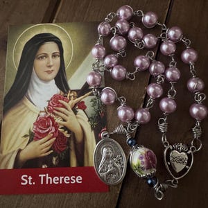 May include: A pink pearl rosary with silver wire and a silver heart charm. The rosary has a silver medal with an image of Saint Therese of Lisieux. The card in the background reads "St. Therese".