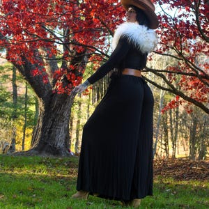 May include: A person wearing a brown cowboy hat, a white fur collar, and a black jumpsuit with a brown belt. The background features a vibrant red tree and green grass.