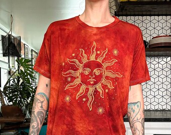 May include: A red tie-dye t-shirt with a gold sun and stars design. The sun has a face and is surrounded by small stars.