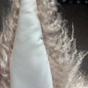 May include: A close-up of the inside of a light pink fluffy jacket. The lining is white satin with a small metal hook and loop closure.