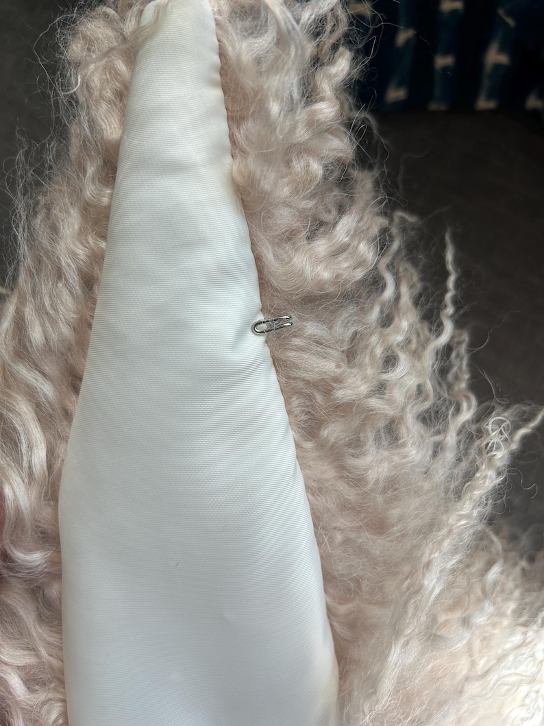 May include: A close-up of the inside of a light pink fluffy jacket. The lining is white satin with a small metal hook and loop closure.