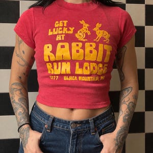 Könnte beinhalten: Ein rotes, kurzärmeliges bauchfreies Oberteil mit gelben Grafiken von zwei Hasen und dem Text "GET LUCKY AT RABBIT RUN LODGE 1977 BLACK MOUNTAIN, NC". Das Shirt hat einen Rundhalsausschnitt und eine körpernahe Passform, kombiniert mit einer blauen Jeans.