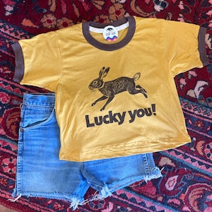 May include: A mustard yellow ringer t-shirt with dark brown trim at the neck and sleeves. The front features a black graphic of a running rabbit and the text "Lucky you!". Below the shirt are blue denim shorts with a frayed, raw-edge hemline.