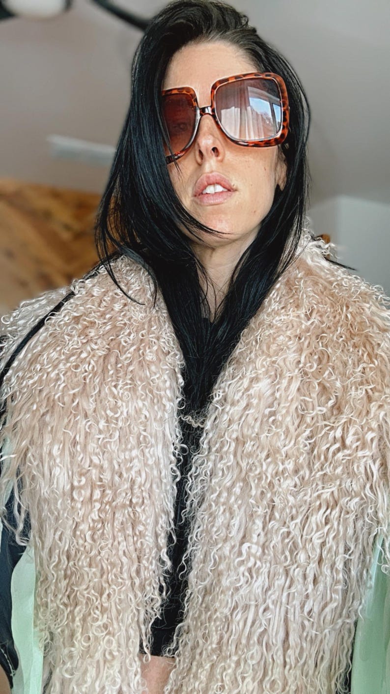May include: A woman wearing a light brown curly fur vest with a black top underneath. She is wearing large, brown, tortoise shell sunglasses.