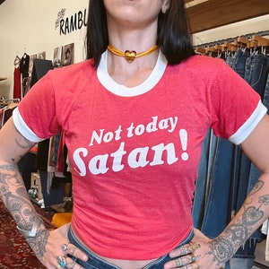 K&ouml;nnte beinhalten: Rotes Ringert-Shirt mit wei&szlig;em Besatz und dem Text "Not today Satan!" in Wei&szlig;. Das Model tr&auml;gt das Shirt mit einer goldenen Herz-Chokerkette.