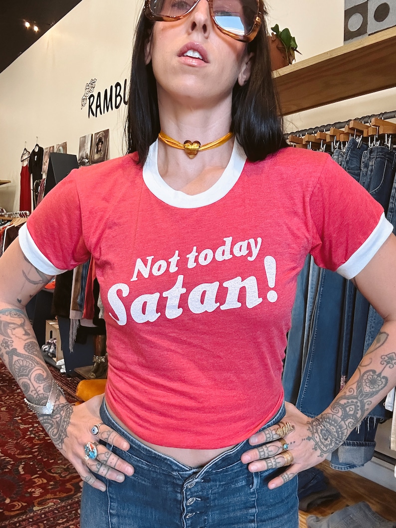 K&ouml;nnte beinhalten: Rotes Ringert-Shirt mit wei&szlig;em Besatz und dem Text "Not today Satan!" in Wei&szlig;. Das Model tr&auml;gt das Shirt mit einer goldenen Herz-Chokerkette.