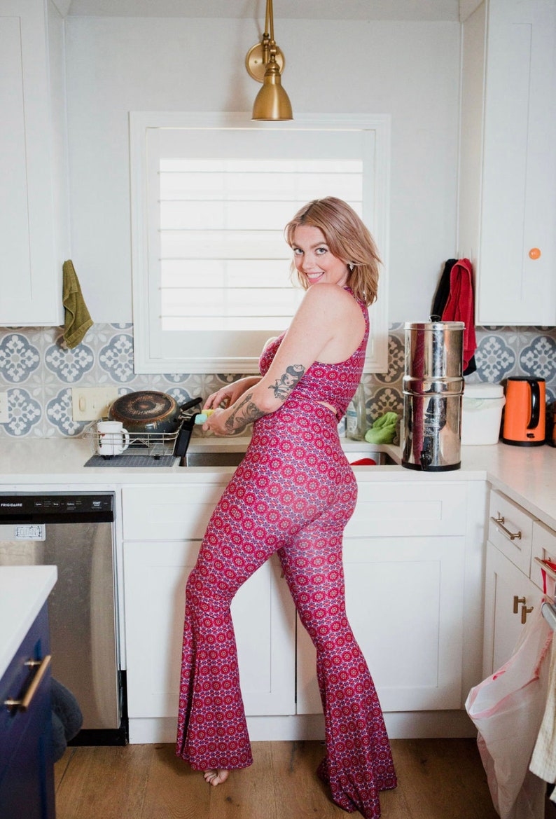 May include: A woman wearing a red and blue patterned pantsuit with a flared leg style. She is standing in a kitchen with white cabinets and a stainless steel dishwasher.